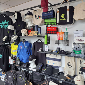 Retail display wall of branded merchandise, including caps, tote bags, drinkware, apparel, and backpacks with various logos.